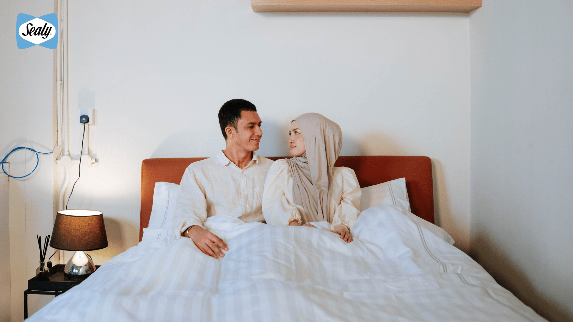 Why Is Quality Sleep Important During Ramadan? 3 Couple sitting in bed, looking at each other with white bedding in a bright bedroom.